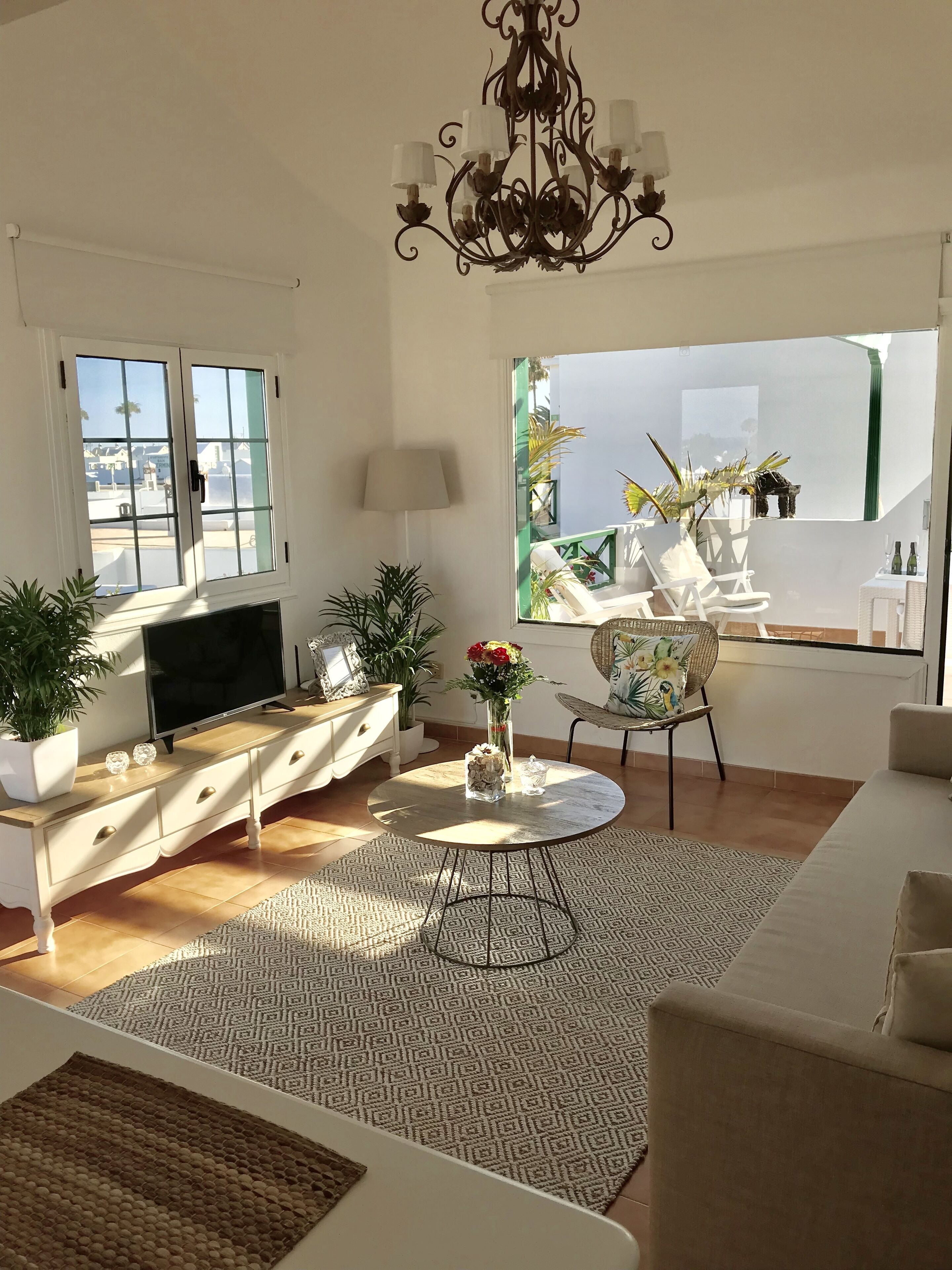 Un salon spacieux avec un téléviseur, un canapé et un coin repas avec des chaises. On y trouve également des plantes en pot et un lustre.