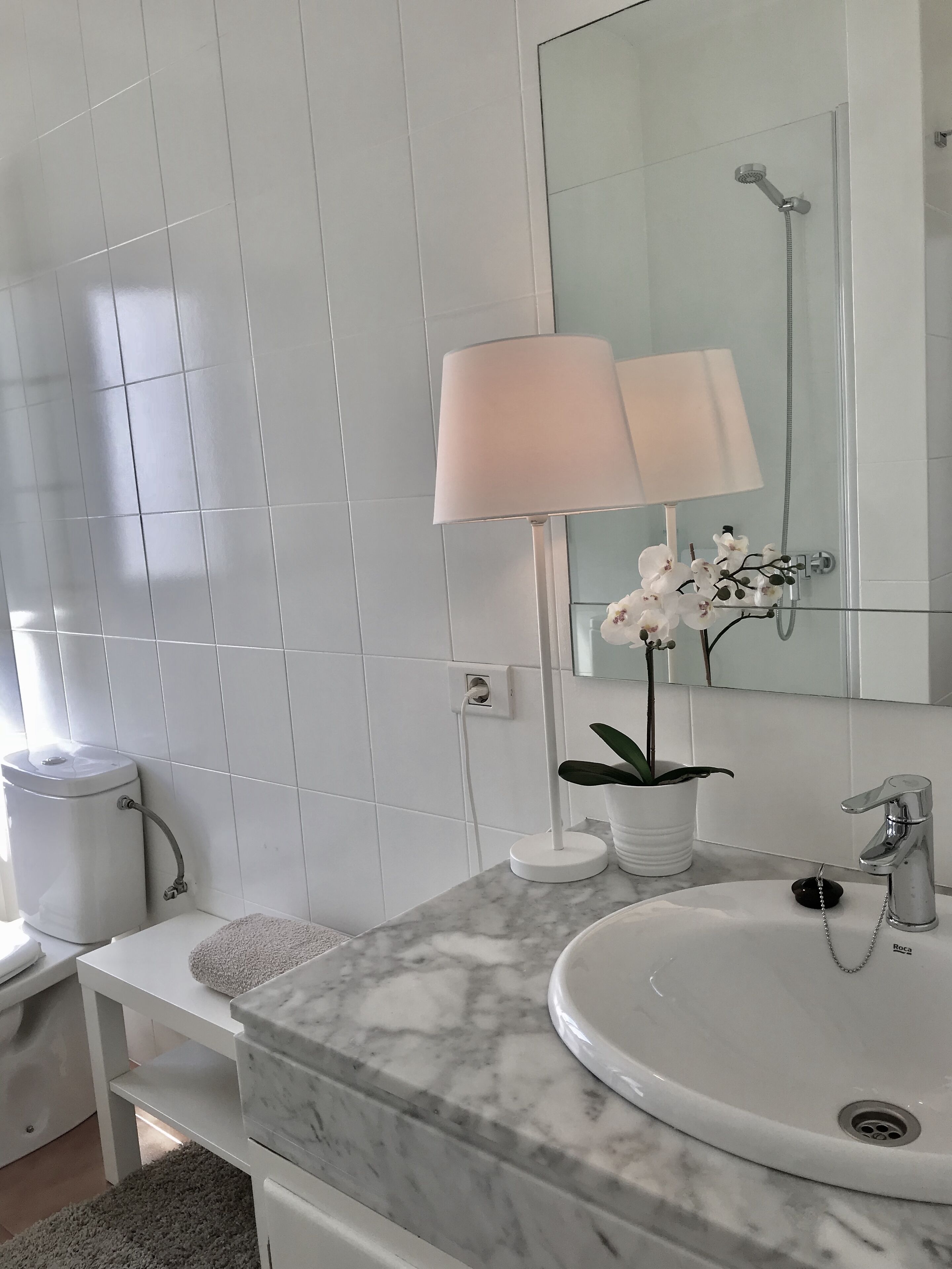 Un baño con lavamanos blanco, encimera de mármol, espejo, ducha, inodoro y una pequeña planta en maceta.