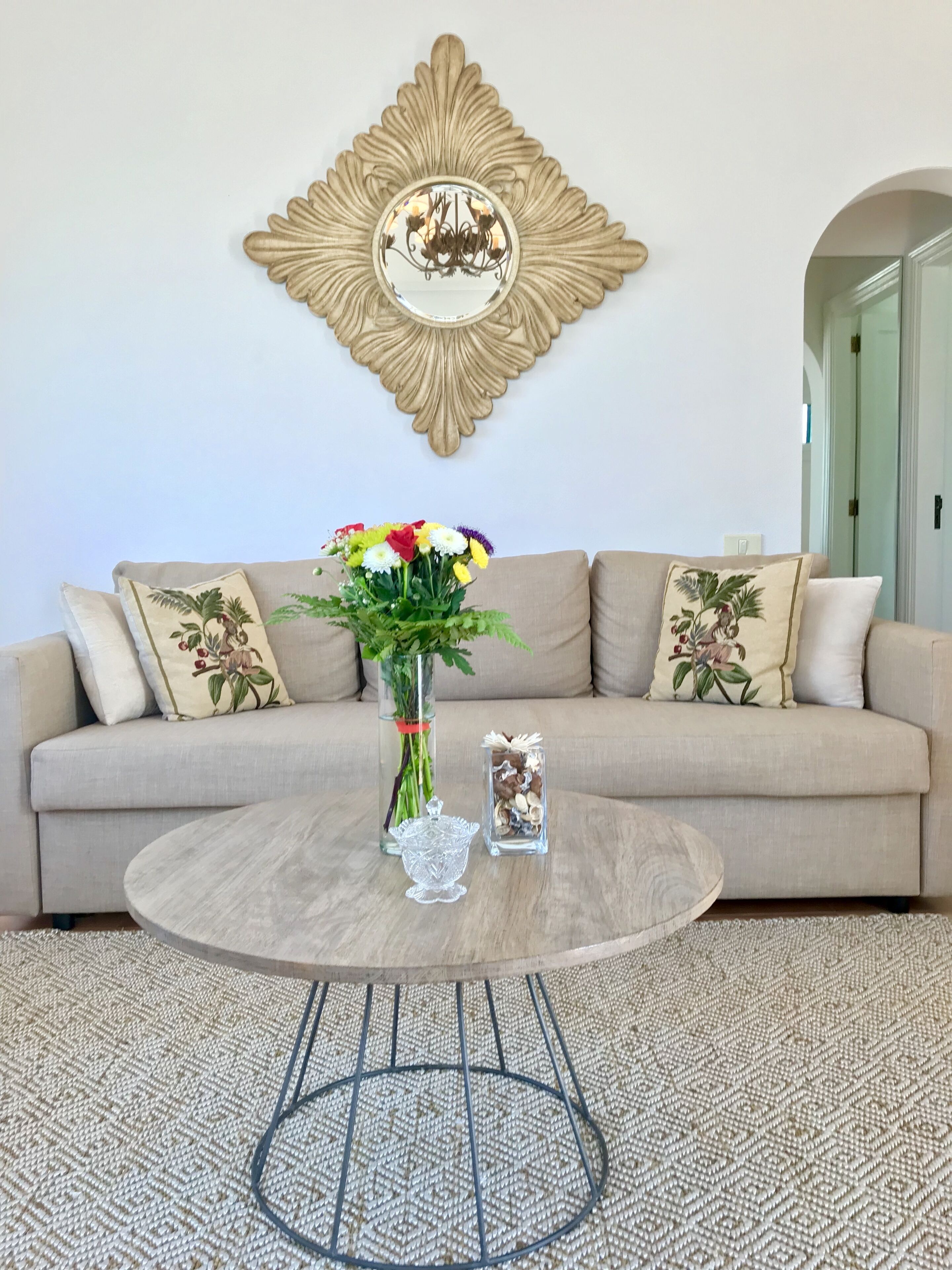 A living room with a beige sofa, a round coffee table, a decorative mirror, and a vase with flowers.