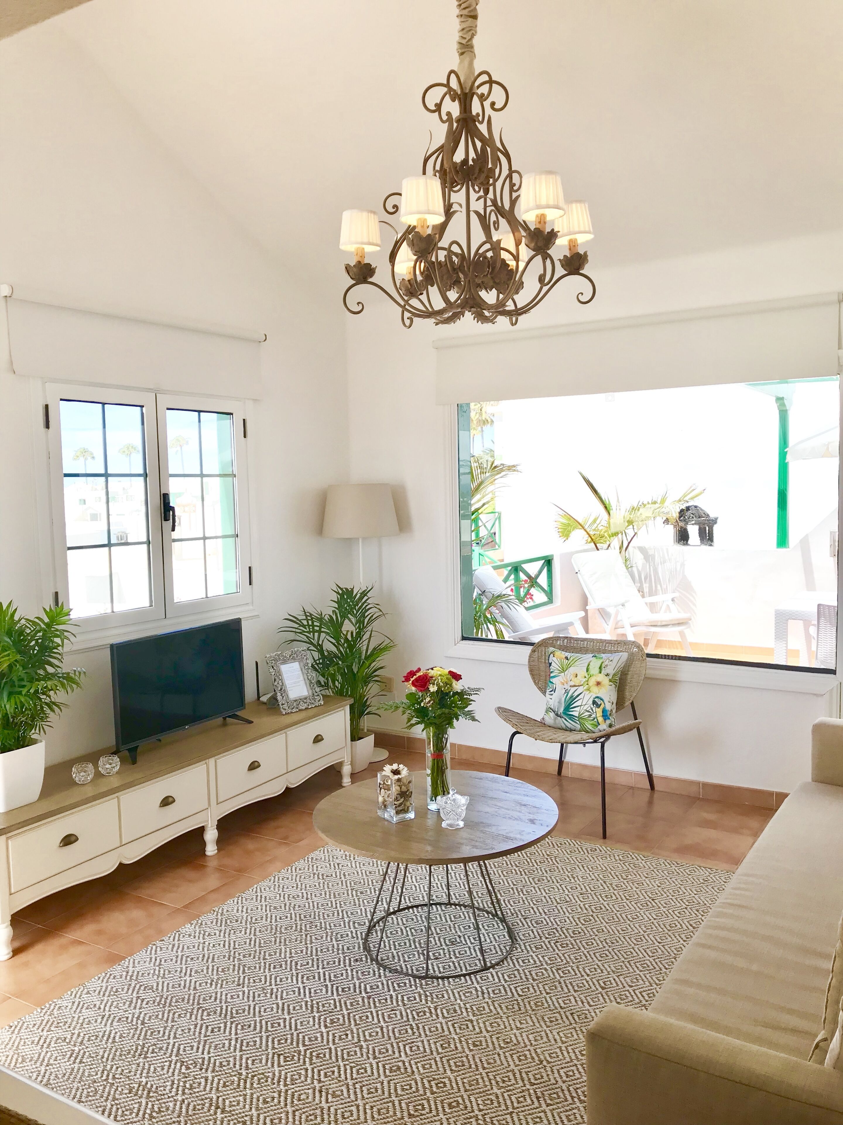 A living room with a TV, white furniture, a chandelier, a window, and a view of the outside.