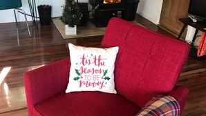 Ein rotes Sofa mit festlichem Kissen, ein schwarzer Kaminofen und ein kleiner Weihnachtsbaum.