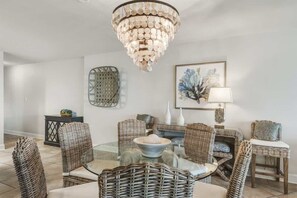 Uma sala de jantar com mobiliário de vime, uma mesa de vidro, um candelabro e uma pintura abstrata emoldurada.