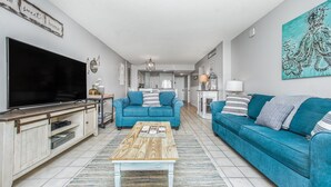 Sala de estar com televisão, sofás azuis, uma mesa de centro e cozinha ao fundo.