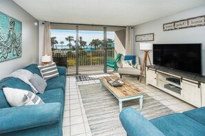 A living room with a teal sofa, a coffee table, a TV, and a balcony with a view of palm trees and the ocean. - AWESOME VIEWS! NEW UPGRADES! (Panama City Beach)