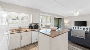 A modern kitchen with white cabinets, a granite countertop, and a built-in island. There is a large window with blinds, a ceiling fan, and a flat-screen TV on a white cabinet. - Beach-side, beautiful low-rise with uncrowded beach! (Destin)