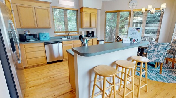 A modern kitchen with wooden cabinets, a central island with stools, a microwave, a coffee maker, and a clock on the wall. - Coastal Vacation Paradise~ Folly Treehouse (Folly Beach)