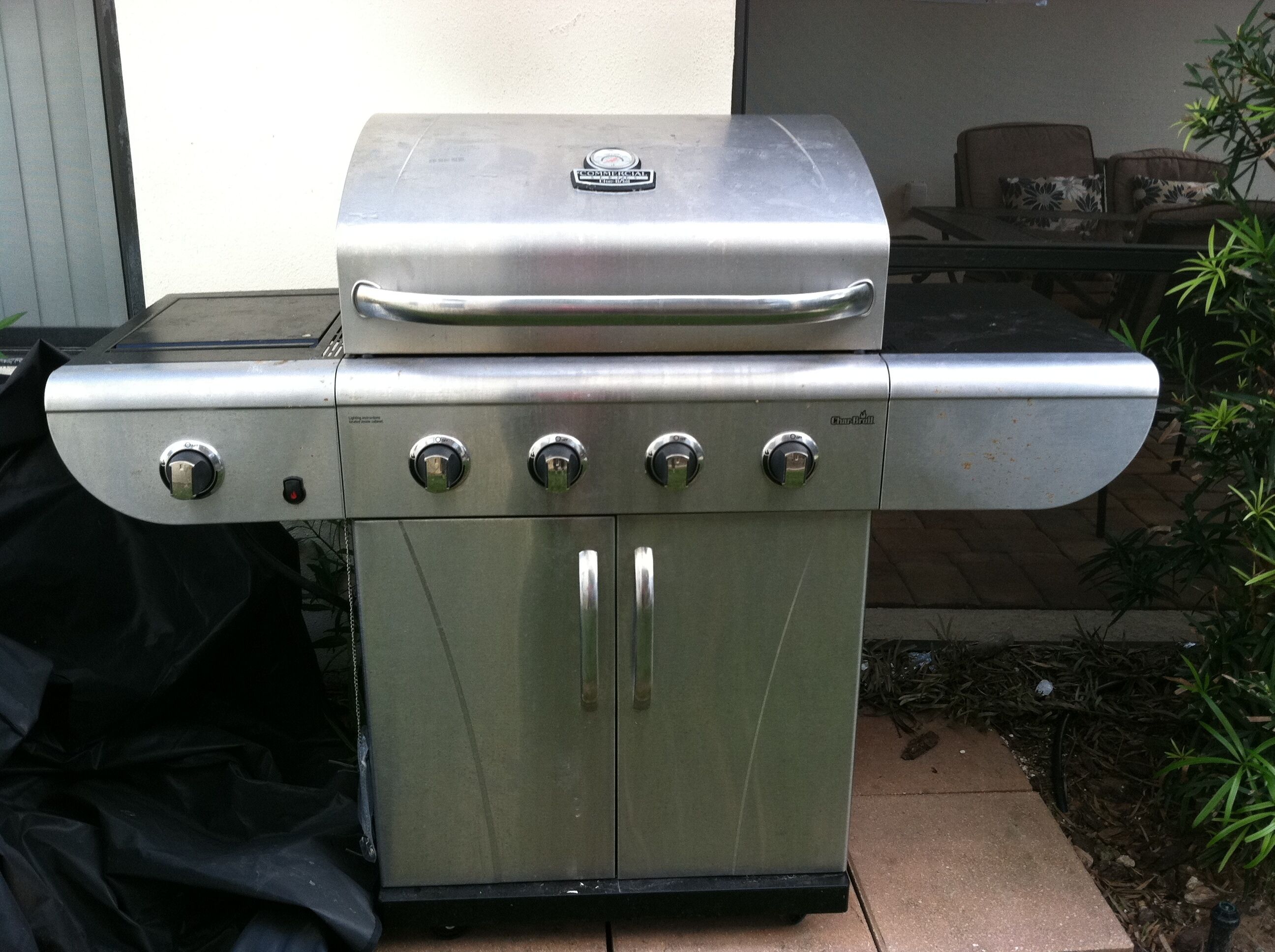 A stainless steel outdoor grill with four control knobs and a lid, placed on a tiled patio.