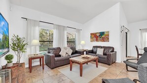 A modern living room with a brown leather sofa, a coffee table, and a grey armchair. There is a painting on the wall and a window with curtains. - 6 miles to Disney: Clean Family 4 Beds, 3.5 Bath, pool/hot tub, game room, grill (Kissimmee)