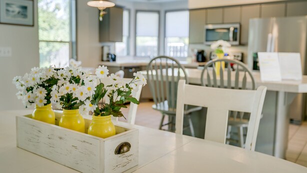 Eine moderne Küche mit weißen Schränken, einem Esstisch mit gelben Vasen voller weißer Blumen und Blick ins Freie durch große Fenster.