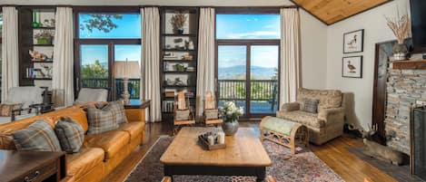 A spacious living room with a wooden ceiling, a large window with a view of the outdoors, a fireplace with a stone surround, and a variety of decorative items on shelves.