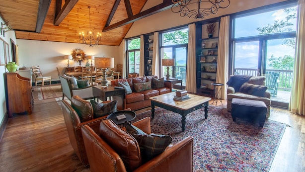 A spacious living room with leather sofas, a wooden coffee table, and a chandelier. Large windows provide natural light and views of the outdoors. - Large Mountain Home with Multi-Mile Views (Boone)