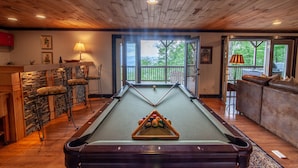 A pool table in a room with wooden ceilings and stone walls, a sofa, and a view of the outdoors. - Large Mountain Home with Multi-Mile Views (Boone)