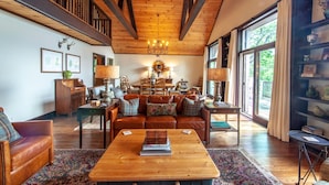A spacious living room with a wooden ceiling, a large window with curtains, a leather sofa, a coffee table, and a bookshelf. - Large Mountain Home with Multi-Mile Views (Boone)