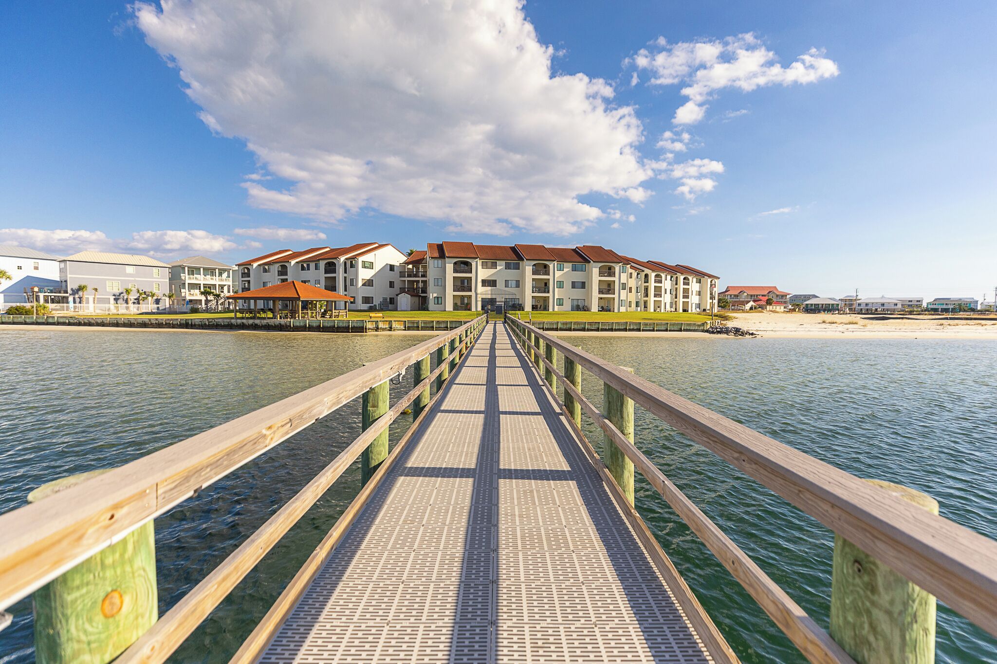 Un muelle de madera que lleva a un hotel frente a la playa, con varios balcones y una vista despejada del océano.