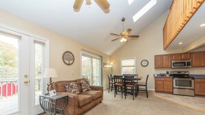 A spacious living area with a brown leather couch, dining table, and chairs, a kitchen with wooden cabinets and stainless steel appliances, and a ceiling fan.