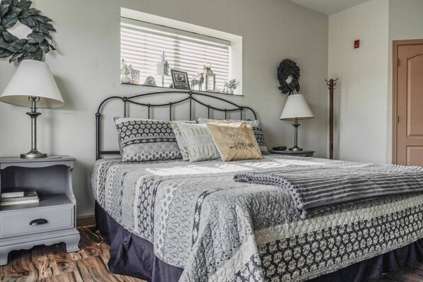 A bedroom with a bed, bedside tables, a window with blinds, a door, and a wall-mounted wreath.