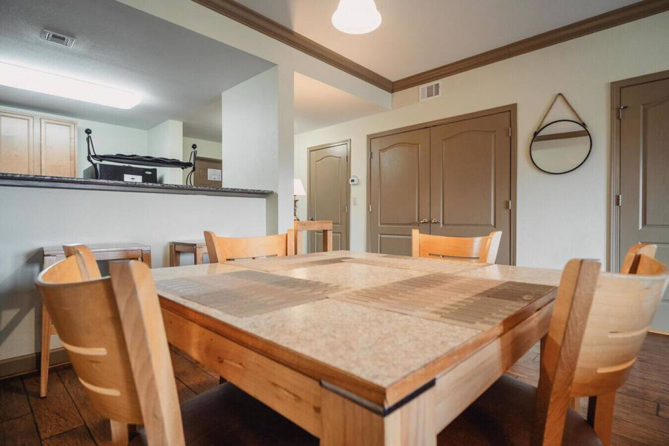 A dining area with a large wooden table and six chairs, a kitchen area with cabinets and a countertop, and a hallway with a door and a mirror on the wall.