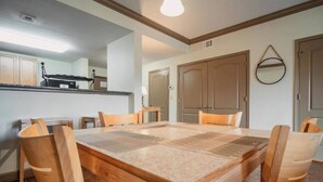 Un espace repas avec une grande table en bois et six chaises, un coin cuisine avec des placards et un plan de travail, et un couloir avec une porte et un miroir au mur.