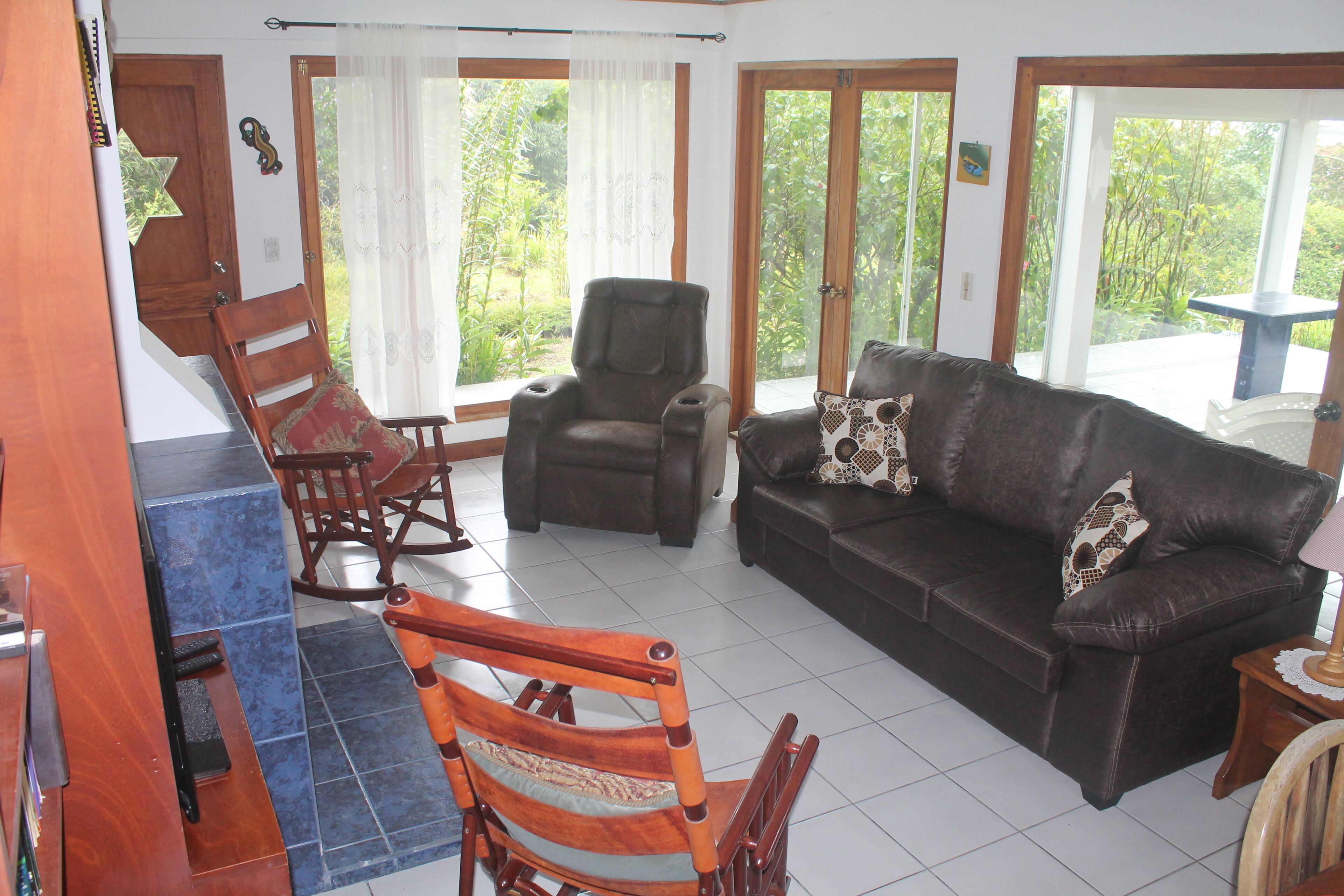 Una sala de estar con un sofá de cuero marrón, una mecedora de madera y un sillón negro. Hay ventanas amplias con cortinas, una mesita pequeña y una silla.