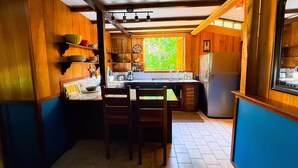 Una cocina con armarios de madera, una mesa de comedor, sillas, un refrigerador y una ventana con vistas a la vegetación.