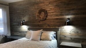 A bedroom with a wooden headboard, a bed with patterned bedding, a nightstand, a wall-mounted lamp, and a decorative wreath.