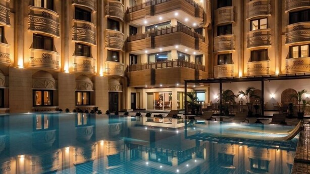 A large indoor swimming pool with a reflection, surrounded by a multi-story building with balconies and ornate architectural details. - Empires Hotel Paradip (Kujang)