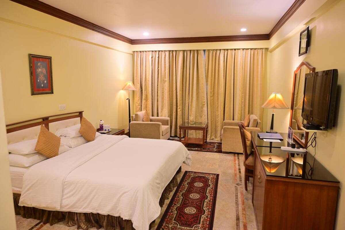 A hotel room with a bed, two chairs, a desk, a television, and a window with curtains.