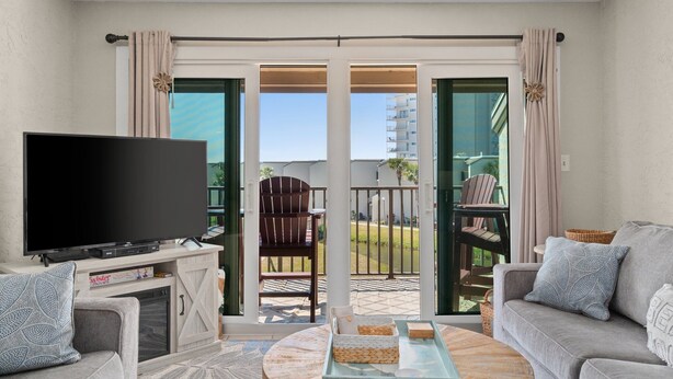 A living room with a flat-screen TV, a sofa, a coffee table, and a balcony with a chair. - Sept 15-19 Reduced! Steps to the Beach & Tootsies! (Panama City Beach)