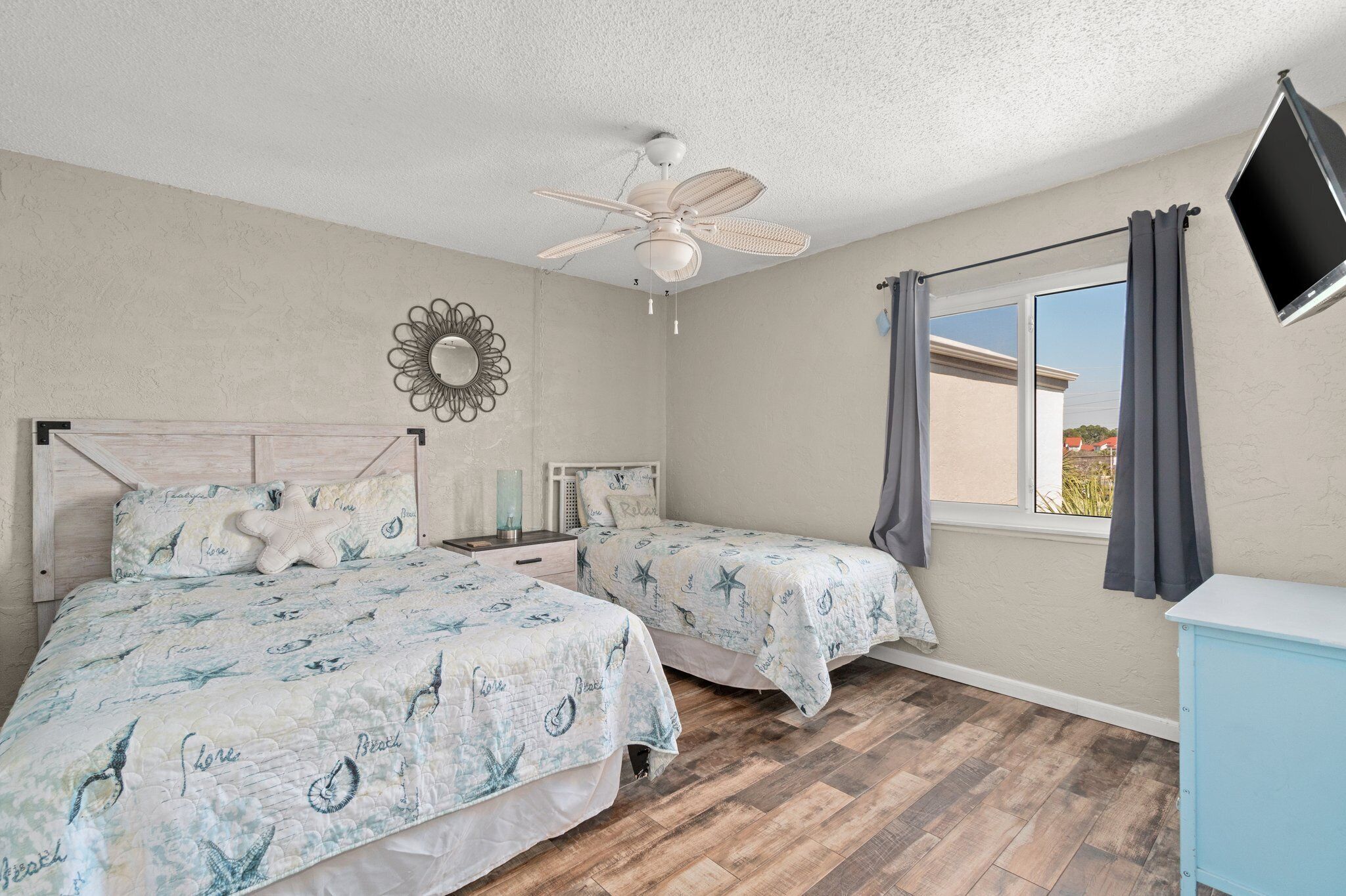 A bedroom with two beds, a ceiling fan, a TV mounted on the wall, and a window with curtains.