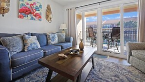 Un salon avec un canapé bleu, une table basse en bois et une vue sur une piscine et des immeubles, visible à travers de grandes portes-fenêtres coulissantes.