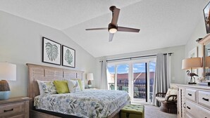 Une chambre à coucher comprenant un lit, des tables de chevet, une commode, un ventilateur de plafond et une grande fenêtre donnant sur des immeubles.