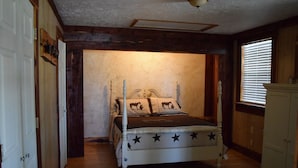 A bedroom with a wooden bed, a cow-themed pillow, a star-patterned bedspread, a cowhide rug, and a window with blinds. - Allure charming peaceful wooded Hill Country like views with easy Lake access (eastland)