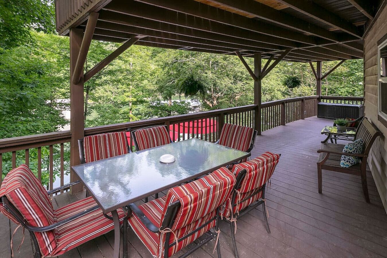 A covered outdoor deck with a glass-top table and striped cushioned chairs, surrounded by a wooden railing and overlooking a lush green landscape.