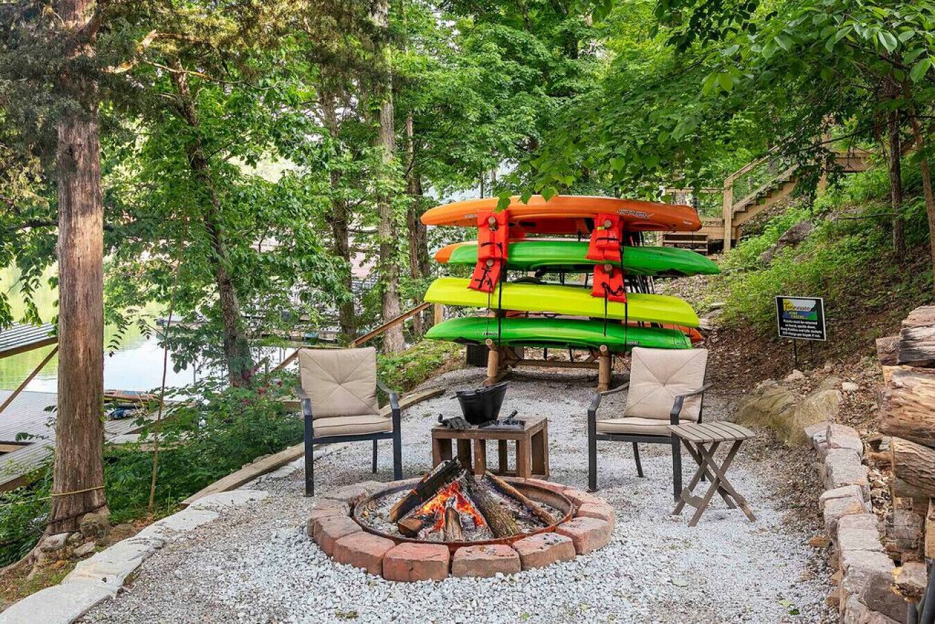 A fire pit surrounded by outdoor seating, kayaks stacked on a wooden platform, and a natural setting with trees and a body of water in the background.