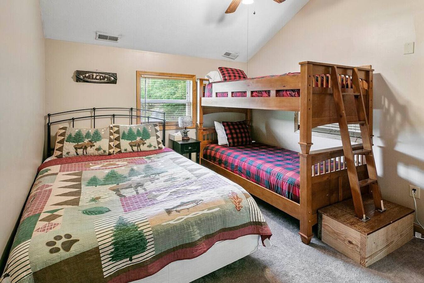 A bedroom with a bunk bed, a bed with a quilt, a 'WELCOME' sign, a window, and a ladder.