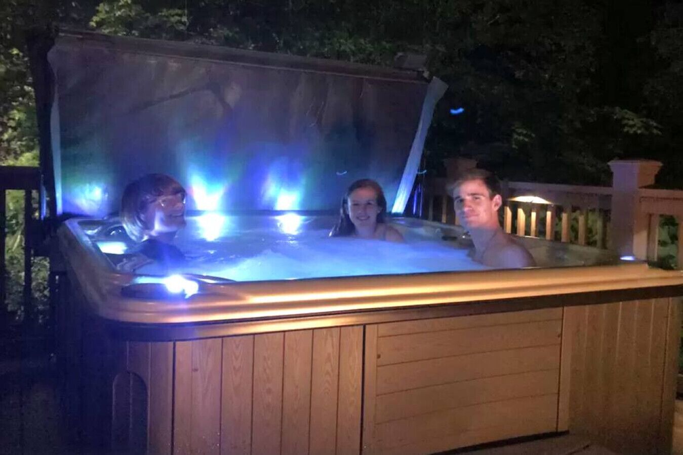 Ein Whirlpool mit drei Personen darin, umgeben von einer Holzterrasse und Bäumen.
