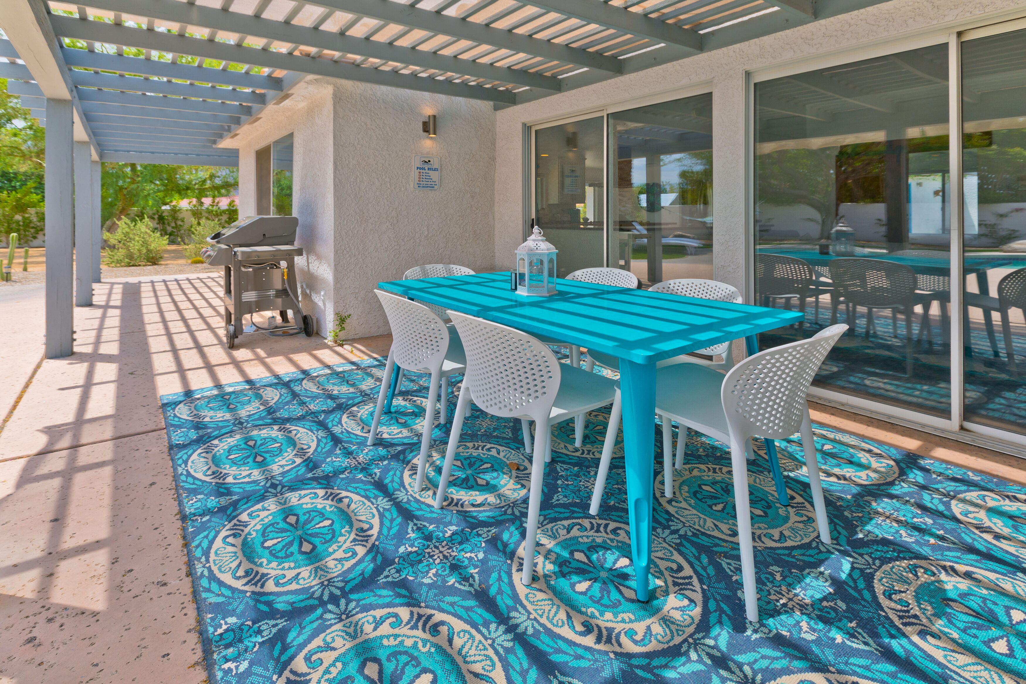 An outdoor patio with a turquoise table and white chairs, a grill, and patterned floor mats.