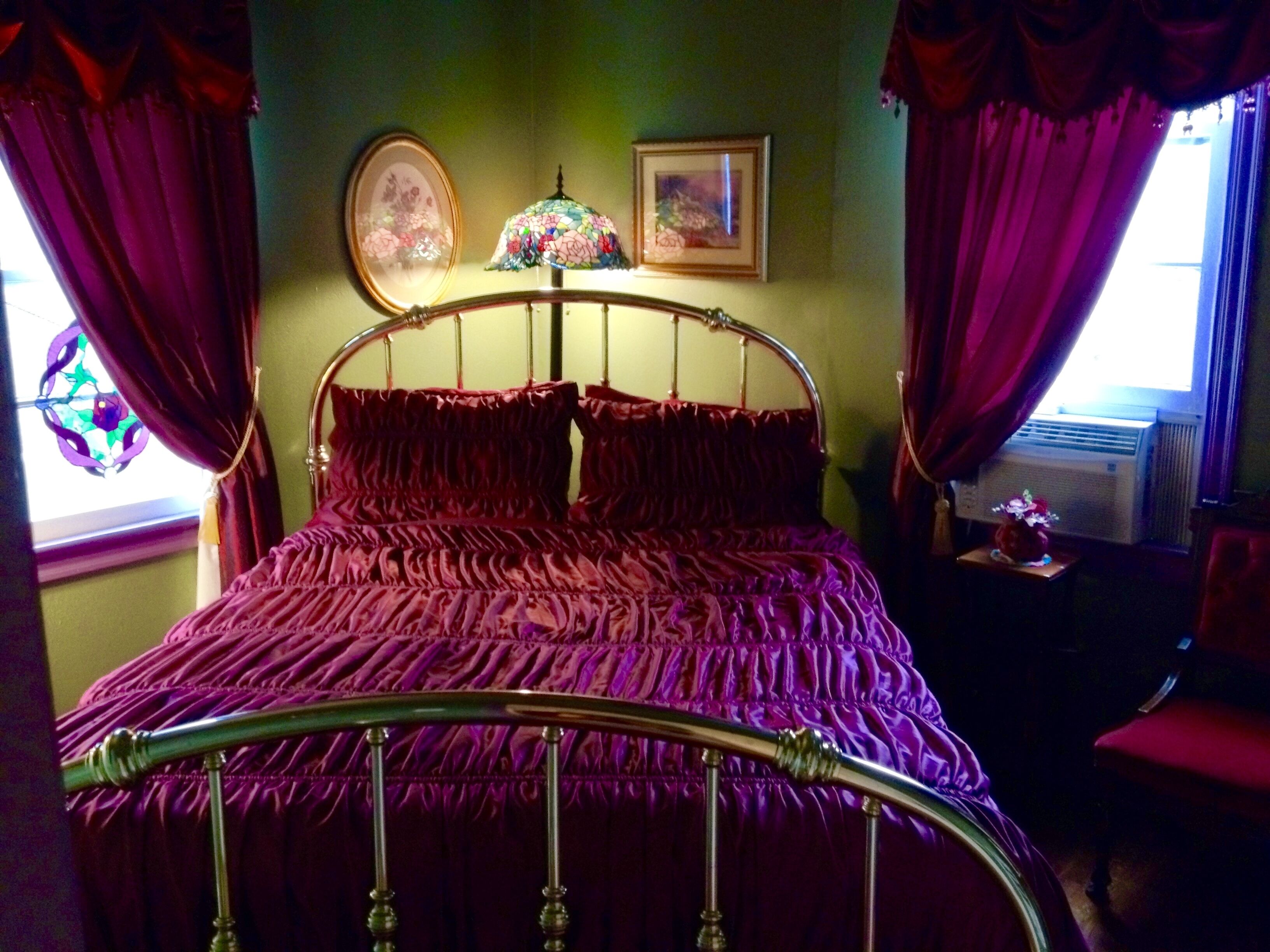 A bed with a metal frame and a purple bedspread in a room with green walls, a window with purple curtains, and a framed picture on the wall.