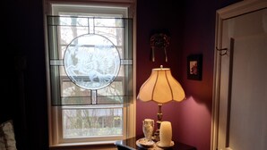 Ein Zimmer mit Fenster, das ein dekoratives Glasmuster zeigt, einem Nachttisch mit Lampe, Kerze und Dekorationsobjekt.