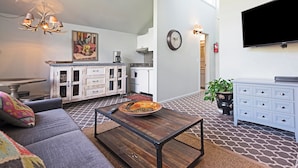 Un salon comprenant un canapé, une table basse et un téléviseur. Il y a également un coin cuisine avec des placards et une horloge au mur.