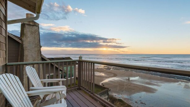 A wooden deck with white Adirondack chairs overlooking a beach with a sunset view.