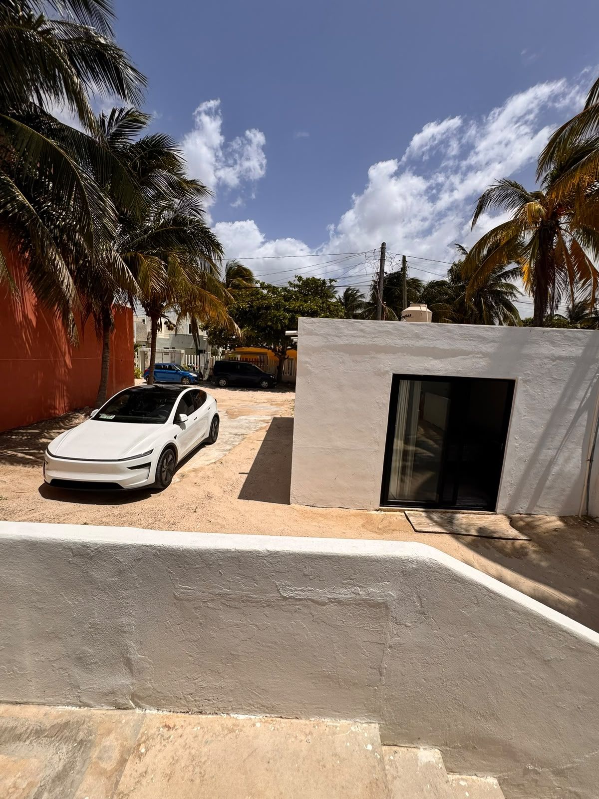 A white car parked in a driveway with a small building and palm trees in the background.