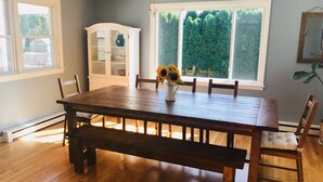 Comedor con mesa de madera, bancos y sillas. Hay un aparador blanco con puertas de cristal y un jarrón con girasoles sobre la mesa.