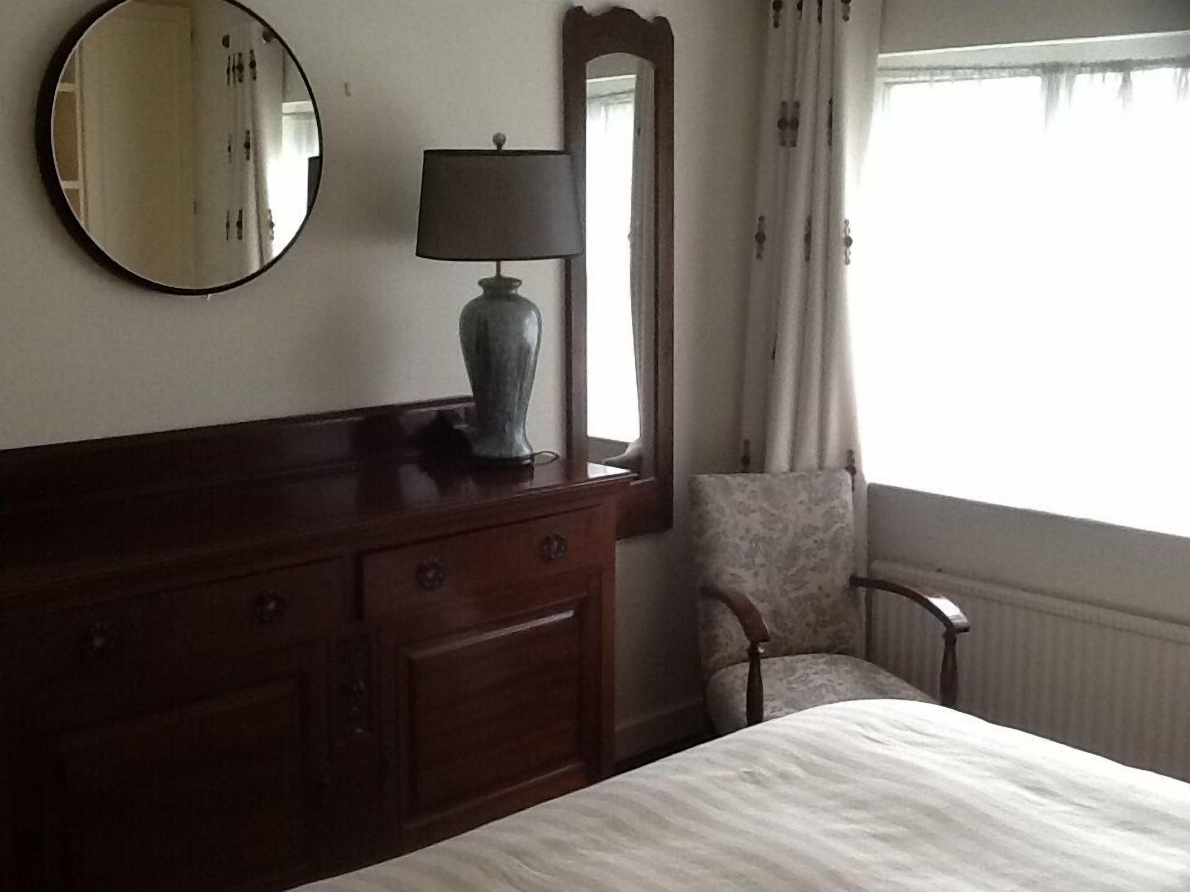 Une chambre d’hôtel avec une commode en bois, un miroir rond, une lampe, une chaise et une fenêtre avec des rideaux.