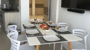 Uma sala de jantar com mesa posta para quatro pessoas, cadeiras brancas, uma televisão de tela plana na parede e uma cozinha ao fundo.