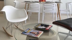 Uma sala de estar moderna com uma mesa de centro de vidro, cadeiras brancas, uma mesa de jantar com frutas e copos, e uma TV de tela plana fixada na parede.
