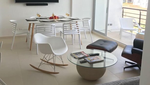 Uma sala moderna com televisão de tela plana, área de jantar com cadeiras brancas, mesa de centro de vidro e sofá cinza.