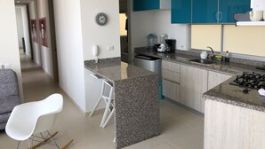Cozinha moderna com bancadas de granito, fogão a gás embutido, cafeteira e liquidificador. Há uma cadeira branca e uma pequena ilha com pia.