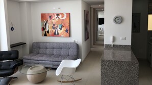 Uma sala de estar moderna com um sofá cinzento, uma cadeira de balanço branca e uma mesa de centro de vidro. Um quadro abstrato e colorido decora a parede, e um relógio está fixado nela.
