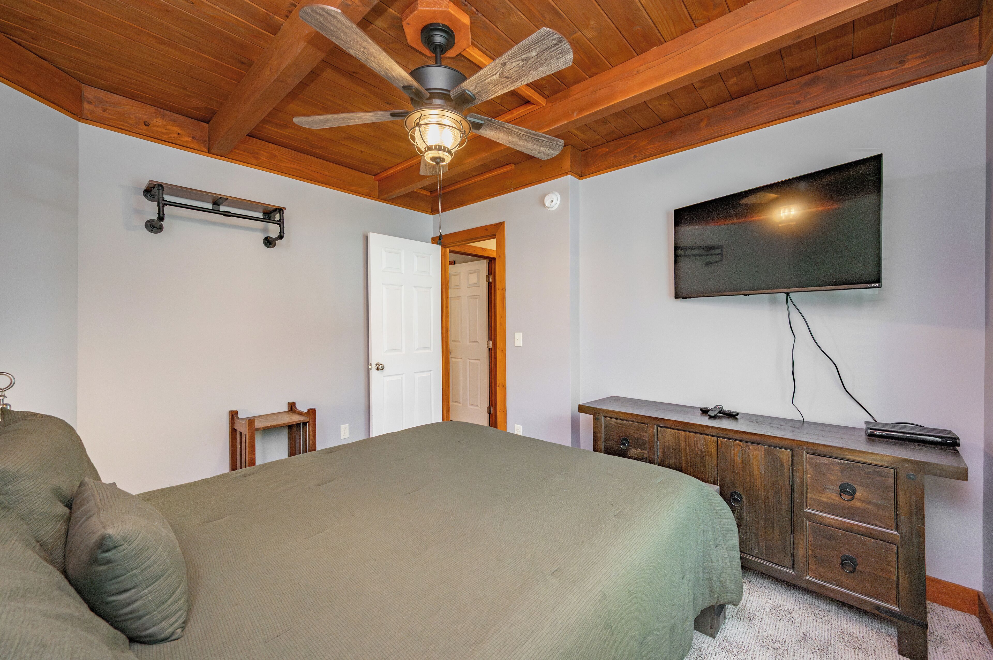 Un dormitorio con cama, ventilador de techo de madera, televisor montado en la pared y cómoda de madera.